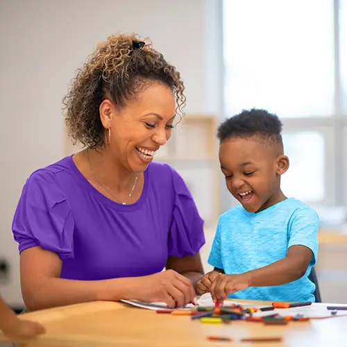 A smiling teacher and a young student laughing together while coloring at a table.