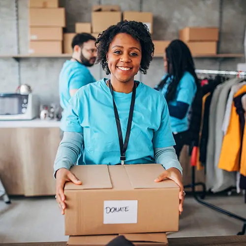 A smiling woman in a blue shirt holds a cardboard box labeled “DONATE” in a room with two other people and shelving.