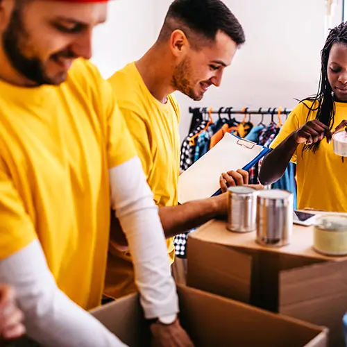 Three volunteers in yellow shirts packing items into cardboard boxes at a food pantry.