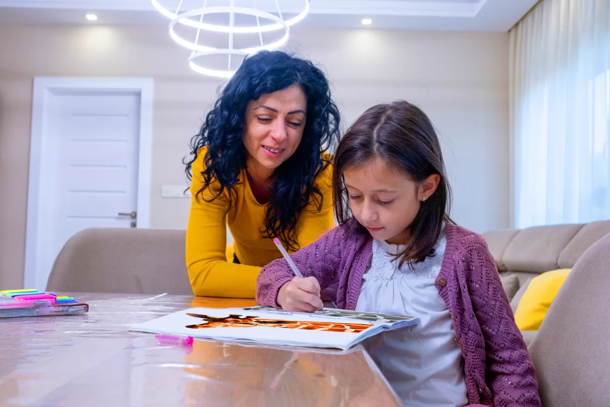 A woman in a yellow top watches a young girl draw in a coloring book at a dining table.
