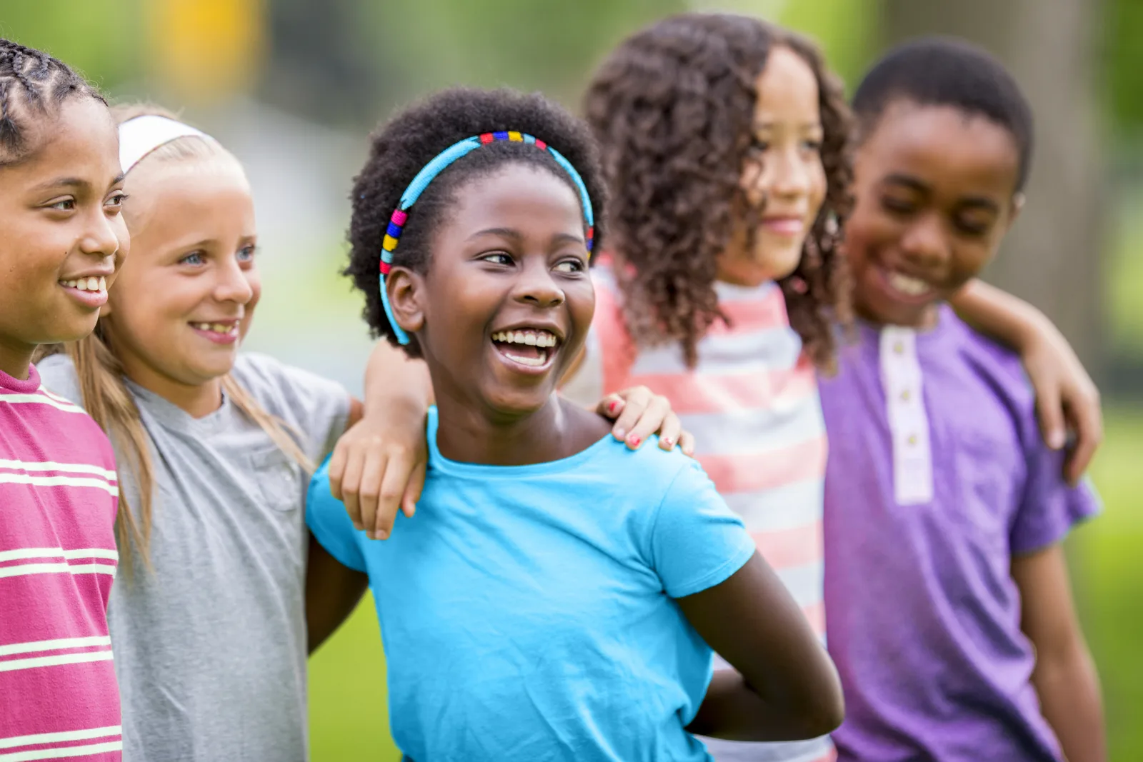 Five children standing in a row with their arms around each other, laughing outdoors.