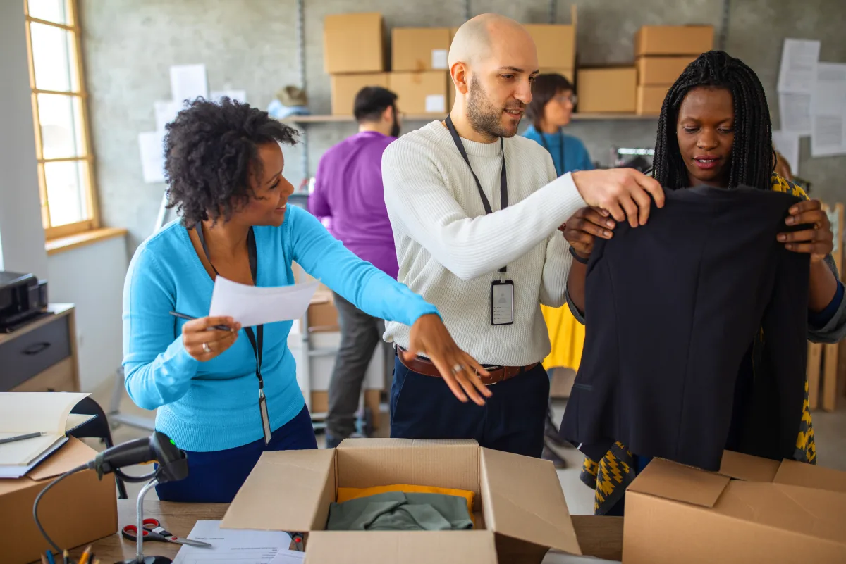 Three people working together to pack clothing into a cardboard shipping box.